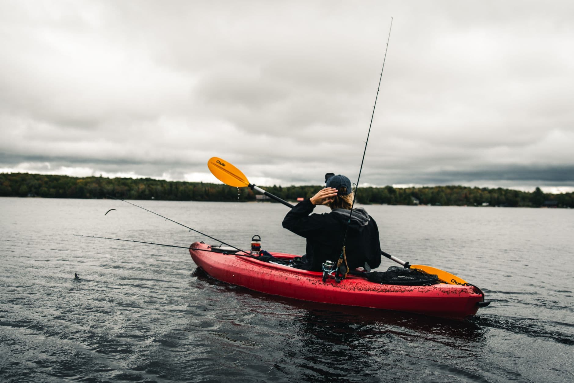 An angler paddling a red fishing kayak rigged with two rods across flat grey water.