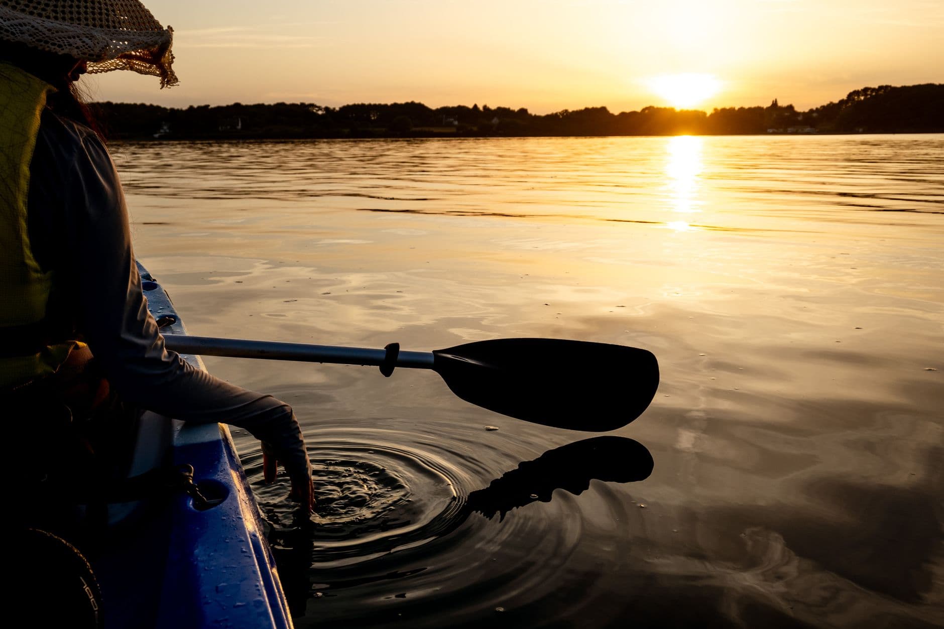 A kayak paddle dipping into calm water at sunset, reflections spreading from the blade.